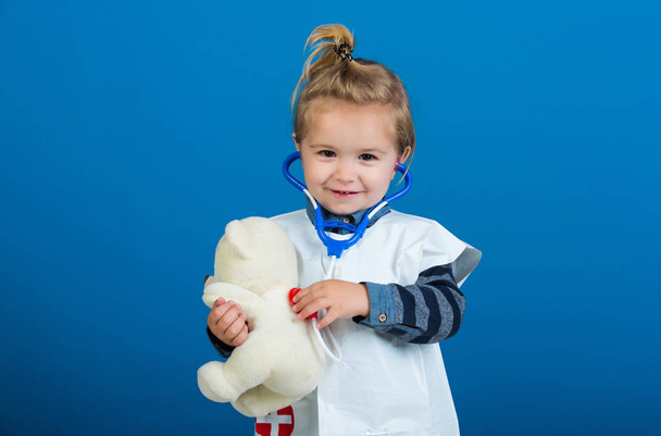 Child veterinarian smile with teddy bear on blue background. Boy in doctor uniform examine toy pet with stethoscope. Veterinary clinic game. Future profession concept, copy space - Fotoğraf, Görsel