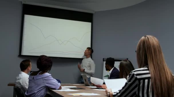 Un joven empleado de la empresa en una camisa blanca informa sobre el progreso del trabajo realizado durante el último año y los planes de desarrollo para el ajetreado trimestre en el diagrama del proyector en el
 - Metraje, vídeo
