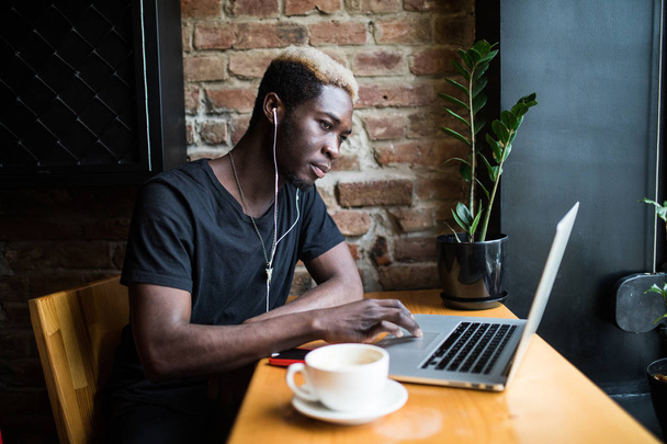 Hombre afroamericano guapo sentado en la cafetería escuchando música y mirando a la computadora portátil
 - Foto, Imagen