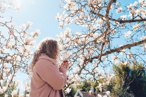 curly-haired girl in a pink coat sniffing a fragrant magnolia under the blue sky in the park - Foto, Bild