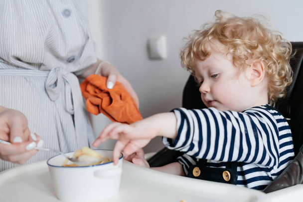 small curly child boy dines in the kitchen in the high chair, useful porridge - Zdjęcie, obraz