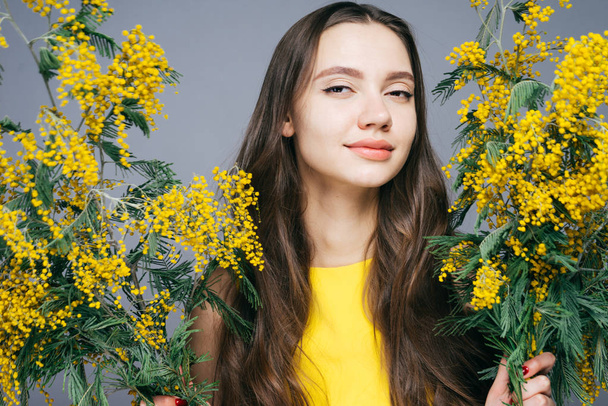 attractive long-haired girl in yellow dress holds a fragrant yellow mimosa, smiling - Foto, immagini
