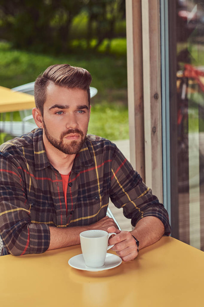 A handsome fashionable male with stylish haircut and beard, wearing fleece shirt, drinking coffee at the cafeteria. - Фото, зображення