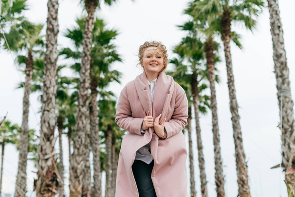 happy young woman in a trendy pink coat walking in the park, enjoying the high green palms and warmth - Foto, Imagem