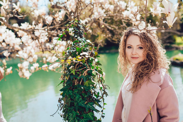 beautiful curly young woman walking in the park, enjoying blossoming trees and a pond, smiling - Фото, изображение
