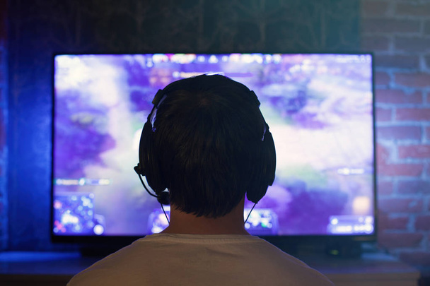 Hráč nebo stuhami v sluchátka s mikrofonem sedí doma v temné místnosti a hraje s kamarády v sítích ve videohrách. Mladý muž sedí u monitoru nebo Tv - Fotografie, Obrázek
