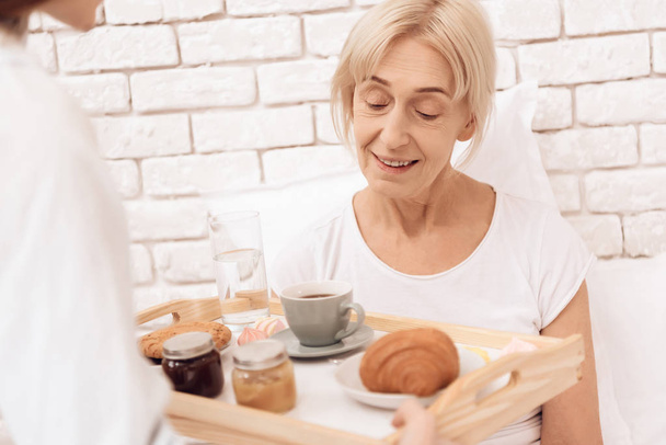 Girl caring for elderly woman and bringing  breakfast coffee and croissant on tray - Photo, Image
