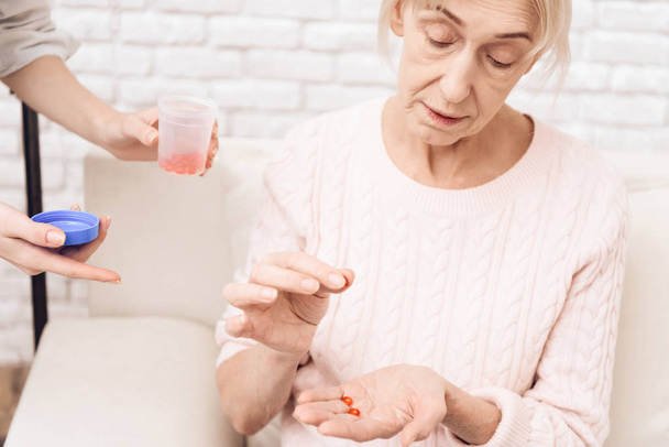 Girl nursing elderly woman on sofa at home and giving woman her pills - Foto, Imagen