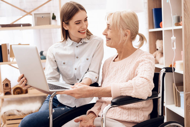 Girl caring for elderly woman in wheelchair and using laptop - Foto, imagen