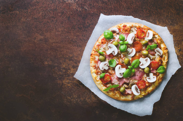 Fresh italian pizza with mushrooms, ham, tomatoes, cheese, olive, basil, on backing paper, dark background, rustic table. Copy space. Homemade with love. Fast delivery. Recipe and menu. Top view - Фото, изображение