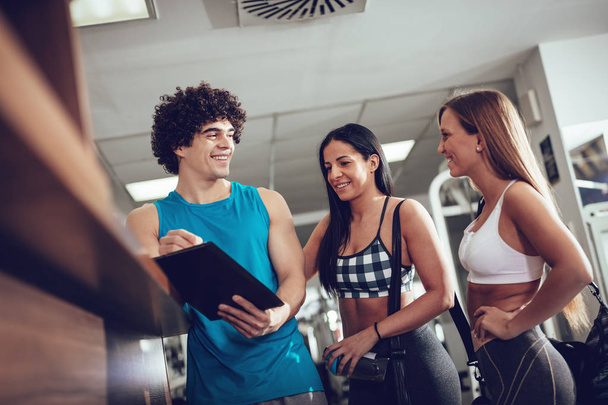 zwei glückliche Freundinnen beraten sich vor dem Training im Fitnessstudio mit einem Fitnesstrainer - Foto, Bild