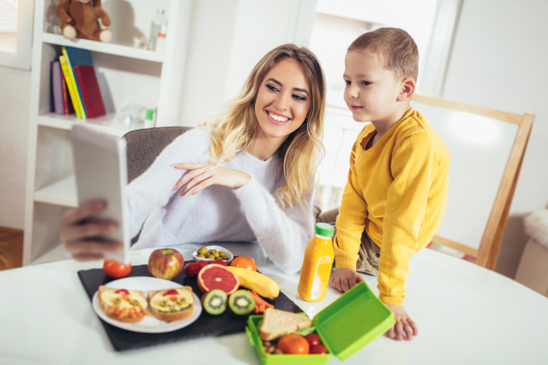 Moeder ontbijt maken voor haar kinderen in de ochtend en een snack voor school, maken een selfie met telefoon - Foto, afbeelding