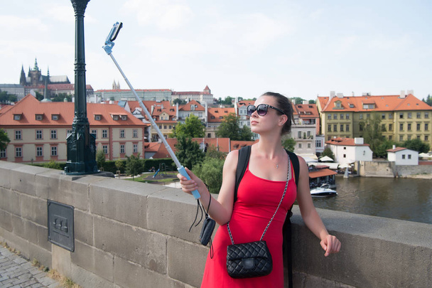 Woman take selfie with mobile phone in prague, czech republic. Woman with smartphone on stick at bridge over river. Girl tourist with fashion look and sensual beauty. Travelling and city sightseeing - 写真・画像