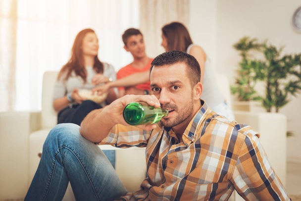 Молодой человек пьет пиво на домашней вечеринке, друзья веселятся на заднем плане
 - Фото, изображение
