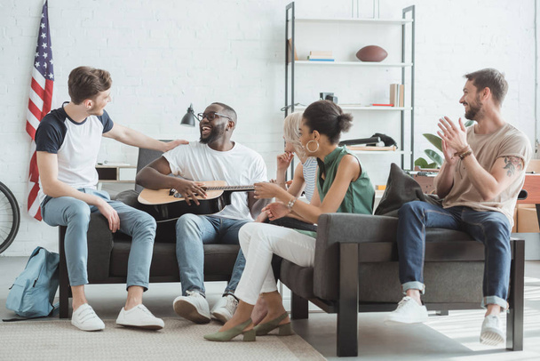 group of multiethnic smiling young people playing guitar in living room - Foto, imagen