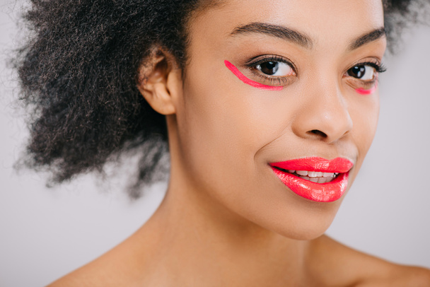 улыбающаяся африканская американка с красными губами и красными мазками краски под глазами, изолированными на сером
 - Фото, изображение