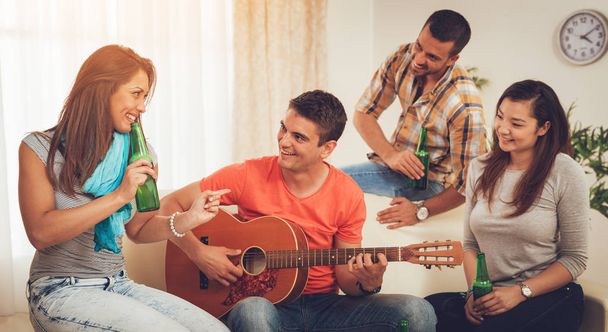 Vier vrolijke vrienden opknoping met gitaar en bier in appartement - Foto, afbeelding