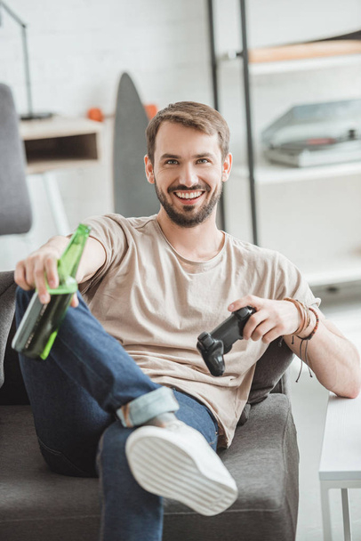 smiling young man sitting with beer bottle and joystick  - Photo, Image