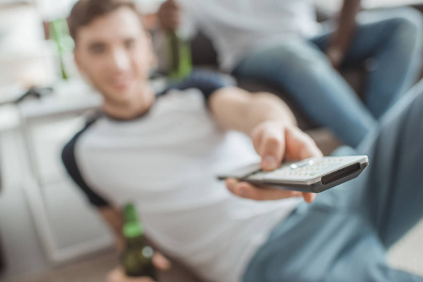 closeup shot of remote in hand of man sitting with bottle of beer  - Foto, imagen
