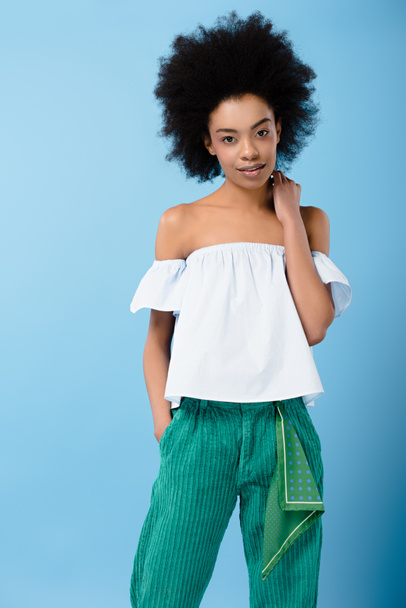 smiling young woman in stylish off-the-shoulder top looking at camera isolated on blue - Foto, imagen