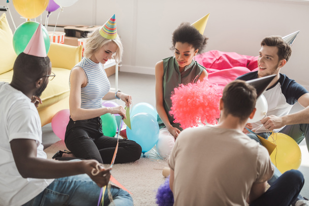 группа молодых мультикультурных друзей в шляпах для вечеринок, сидящих на полу с воздушными шарами в украшенной комнате
  - Фото, изображение