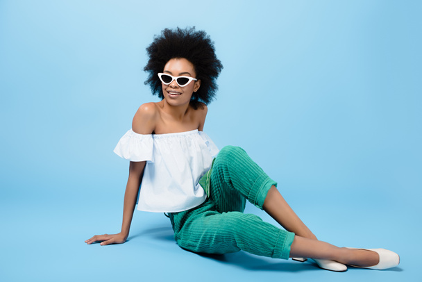 young african american woman in stylish clothes and sunglasses sitting on floor on blue - Photo, Image