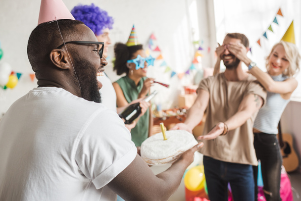 Happy friends covering eyes of young man and greeting him with birthday cake - Photo, Image
