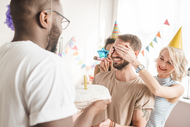 Onnelliset ystävät peittävät nuoren miehen silmät ja tervehtivät häntä syntymäpäiväkakulla
 - Valokuva, kuva