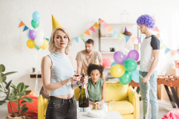 Blonde girl holding champagne bottle in front of partying friends - Photo, Image
