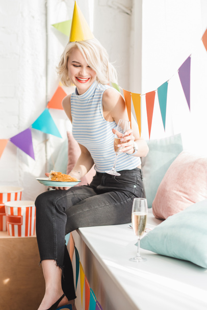 ホーム パーティーでケーキとシャンパン グラスとプレートを保持パーティー ハットで金髪女性を笑顔 - 写真・画像