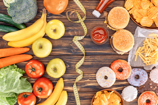 top view of assorted junk food, fresh fruits with vegetables and measuring tape on wooden table    - Photo, Image