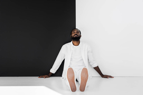 handsome stylish african american man sitting on floor near black and white wall with closed eyes - 写真・画像