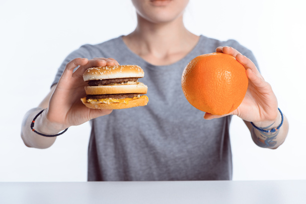tiro cortado de menina segurando hambúrguer e laranja madura
 - Foto, Imagem