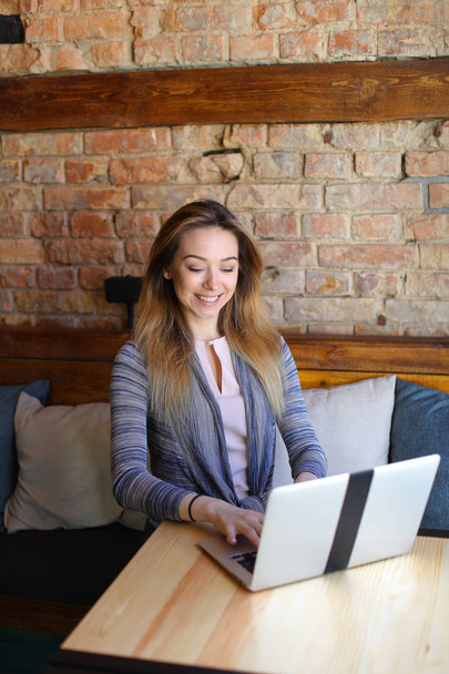 Piękna kobieta siedzi w pobliżu drewniany stół z białym laptopa i kawę w przytulnej kawiarni. - Zdjęcie, obraz