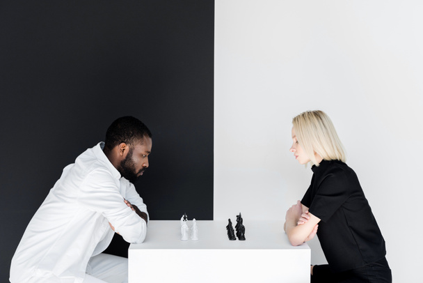 side view of african american boyfriend and blonde girlfriend playing chess near black and white wall - Fotografie, Obrázek