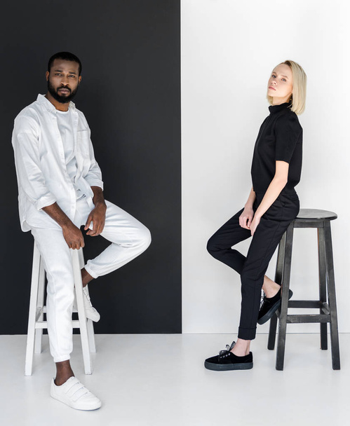 side view of african american boyfriend and blonde girlfriend sitting on chairs near black and white wall - Фото, изображение
