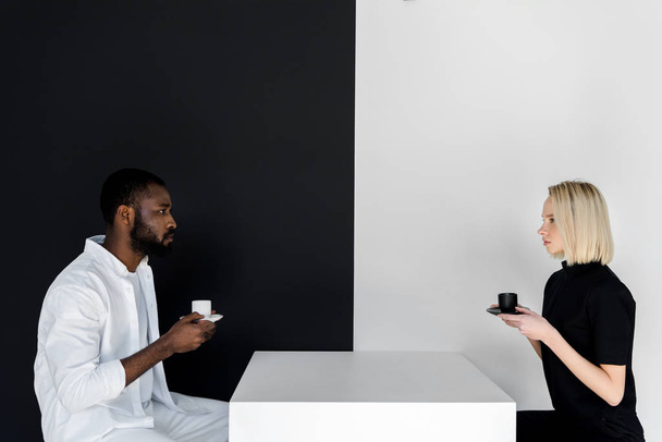 vista lateral de pareja multicultural sentada con tazas de café
 - Foto, Imagen