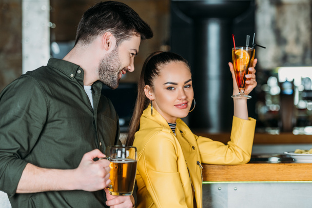 jonge vrouw met cocktail kijken naar camera terwijl man flirten met haar in bar - Foto, afbeelding