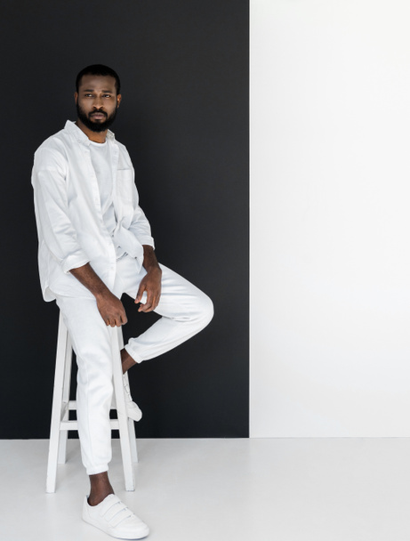 handsome stylish african american man in white clothes sitting on chair near black and white wall - Foto, Imagen