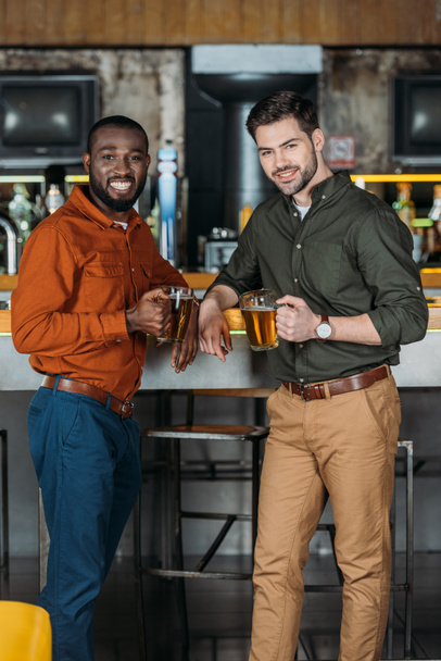 Lächelnde junge Männer mit Bierkrügen am Tresen und Blick in die Kamera - Foto, Bild