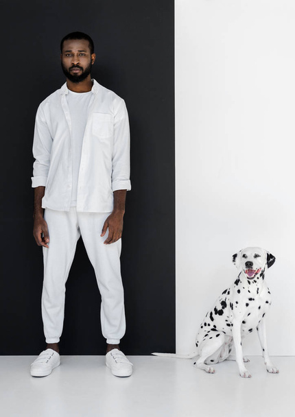 guapo hombre afroamericano con estilo en ropa blanca y perro dálmata de pie cerca de la pared en blanco y negro
 - Foto, Imagen