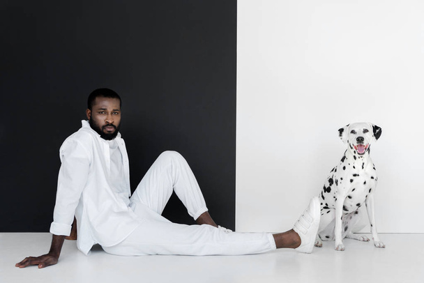 handsome stylish african american man in white clothes and dalmatian dog sitting near black and white wall - Fotografie, Obrázek