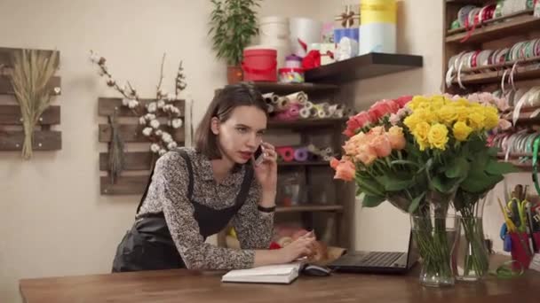Young female worker of floral store is talking by phone and writing in notebook - Footage, Video