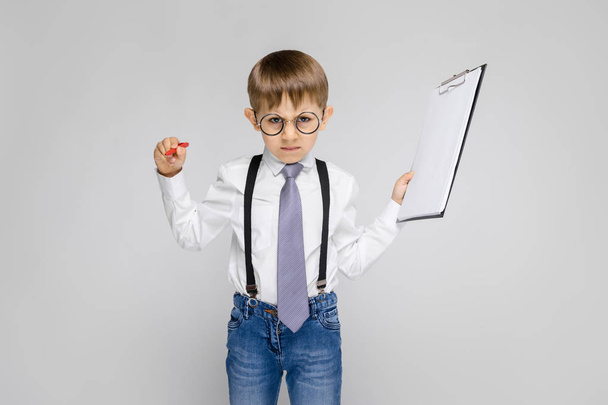 Portrait of adorable serious confident little boy in glasses holding clipboard on grey background - Фото, зображення