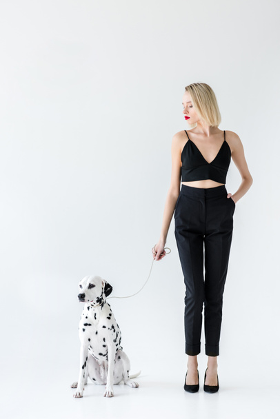 attraktive, stylische blonde Frau in schwarzer Kleidung mit dalmatinischer Hundeleine auf weißem Grund - Foto, Bild