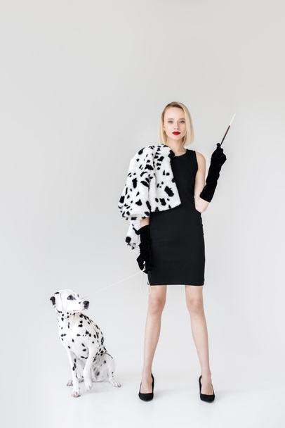attractive stylish blonde woman in black dress holding cigarette, dalmatian dog on floor - Foto, afbeelding