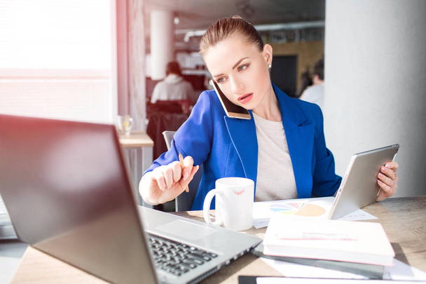 A picture of serious woman working in office. She is holding a tablet in one hand and a pen in the other one. Girl is looking at the laptops screen. Also she has a cup of tea in fromnt of her. - Фото, изображение