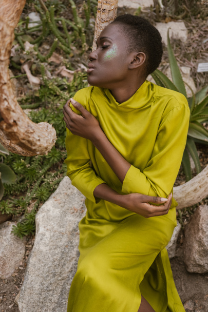tierna mujer afroamericana con los ojos cerrados y maquillaje brillo posando en jardín tropical
 - Foto, Imagen