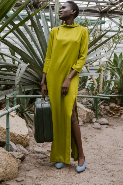 stylová africká americká žena ve žlutých šatech pózuje s cestovní taškou v tropickém skleníku - Fotografie, Obrázek