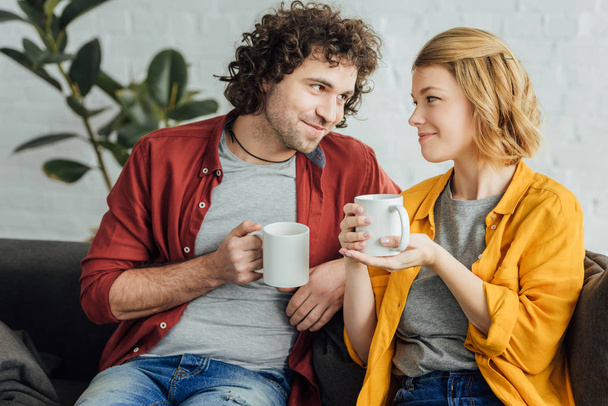 happy young couple holding cups of tea and smiling each other  - Foto, Bild